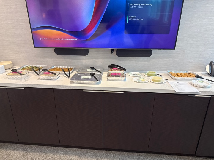Catering trays and serving utensils arranged on a counter beneath a wall-mounted screen.